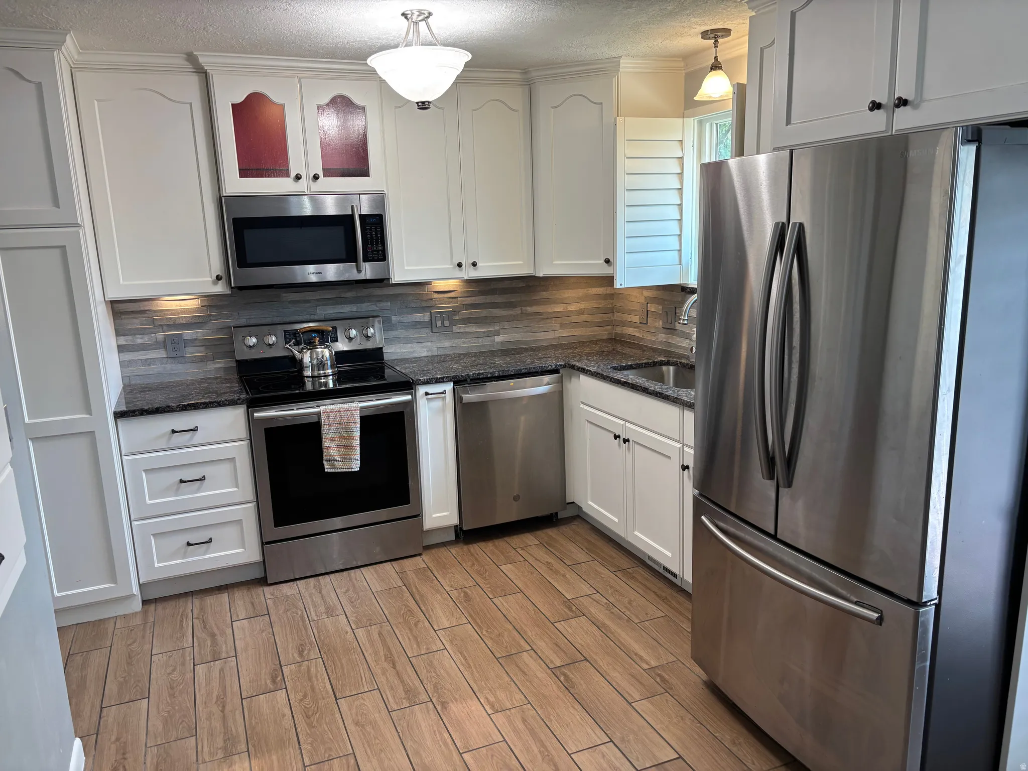 Kitchen featuring white cabinetry with dark hardware, stainless steel appliances, dark speckled countertops, a mosaic tile backsplash, and wood-finish flooring