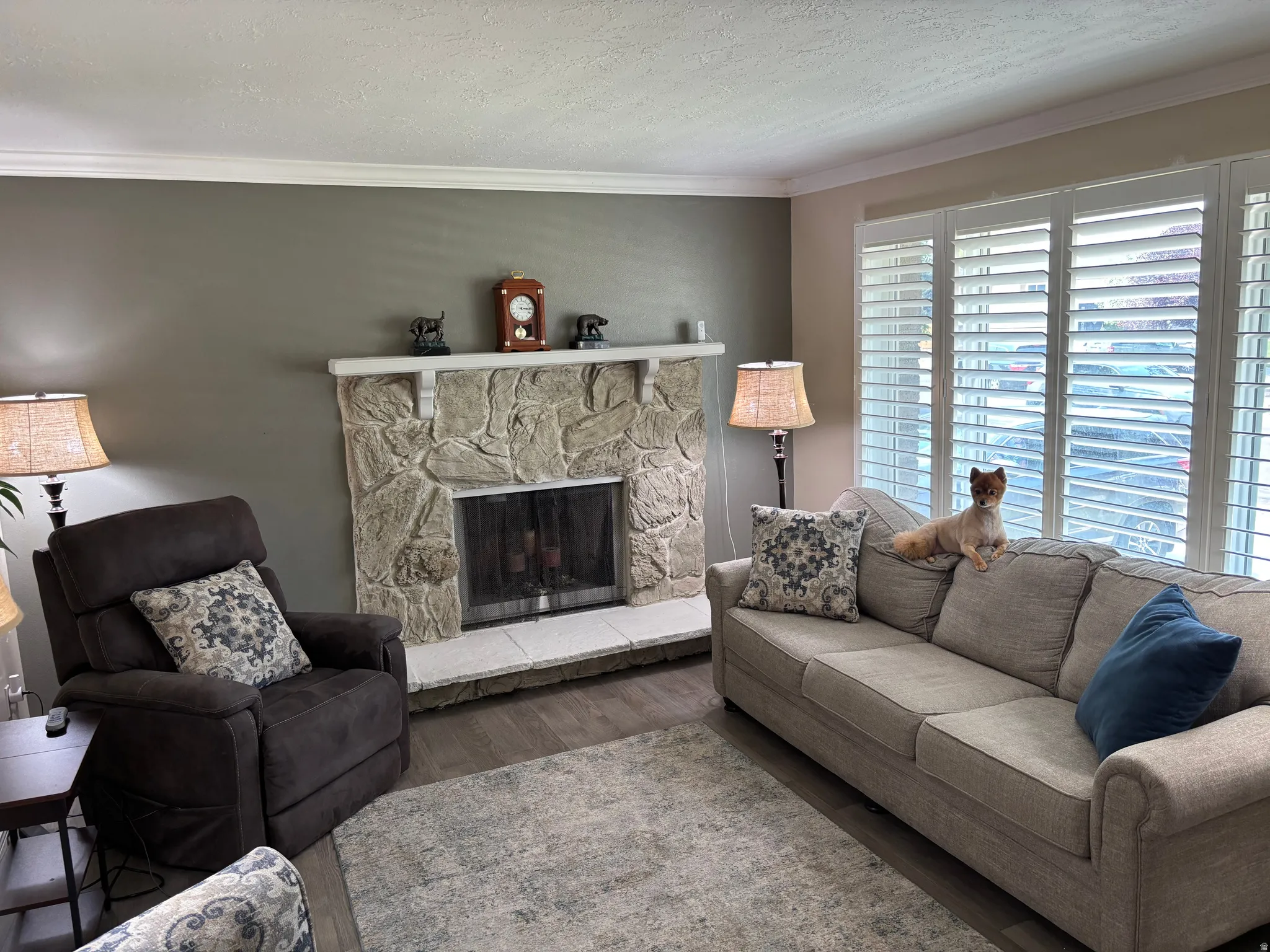 Living room featuring a stone fireplace with a white mantel and hearth, wood-finish flooring, and white plantation shutters on the windows