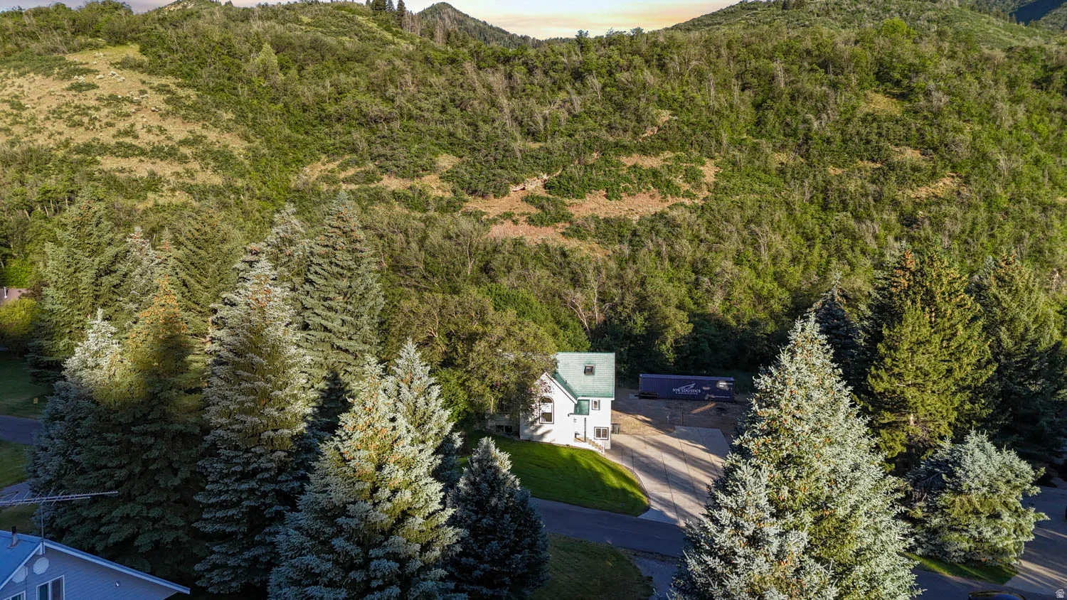 White siding residence with a green roof, set against a backdrop of natural hillsides and surrounded by mature evergreen trees
