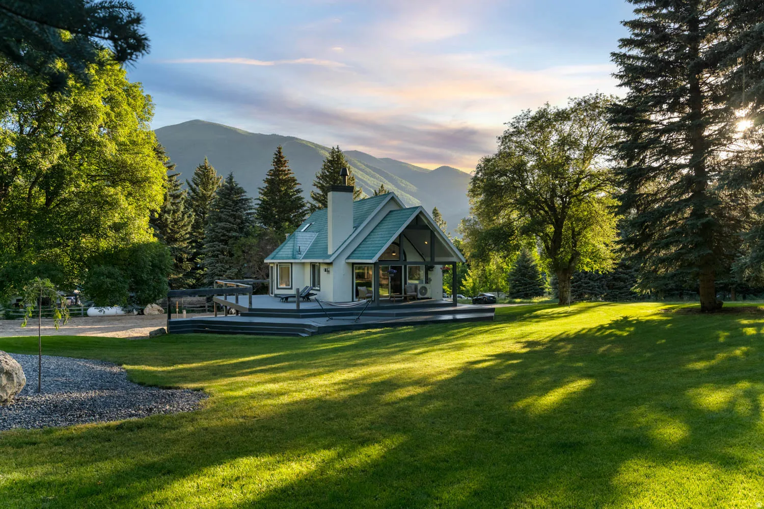 Gable-roofed home featuring a substantial wood deck, expansive lawn, and mature trees, set against a mountainous backdrop