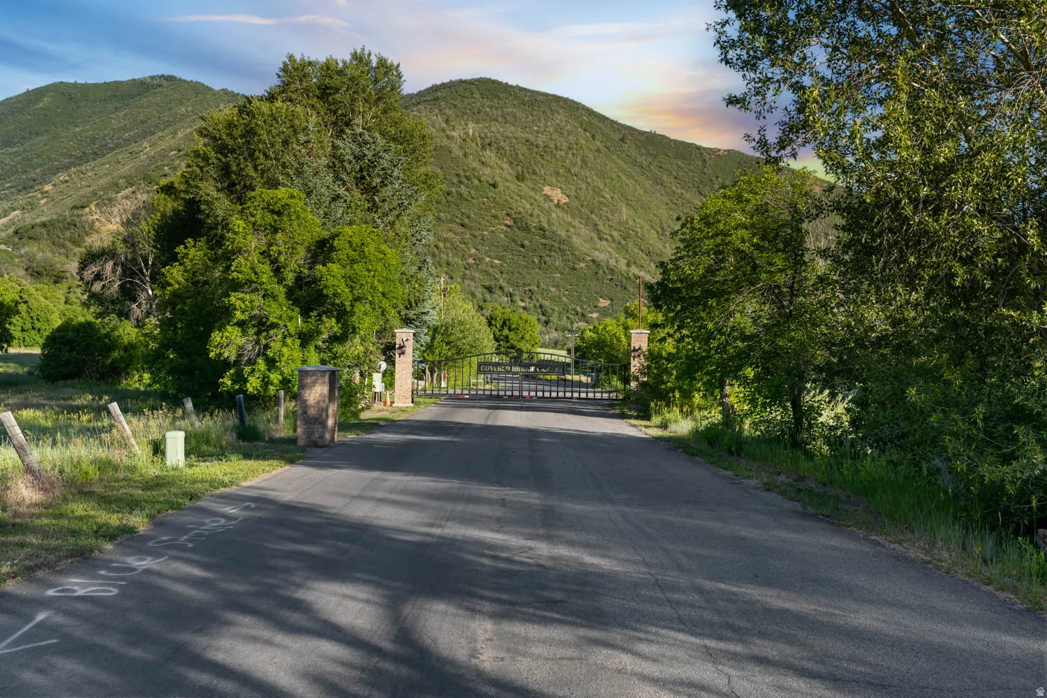 Paved driveway leading to a gated entry, flanked by stone pillars and mature trees, set against a backdrop of rolling, tree-covered hills