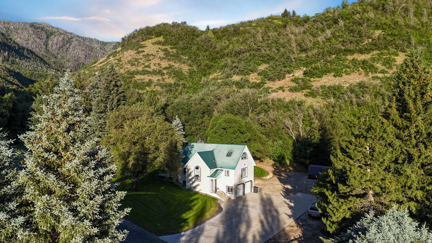 White multi-level residence with a green roof, integrated garage, and arched windows, surrounded by mature trees and verdant mountain terrain