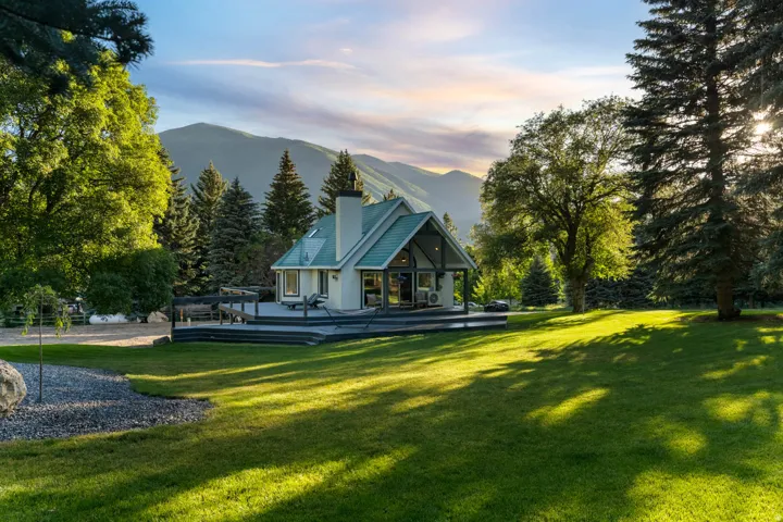 Gable-roofed home featuring a substantial wood deck, expansive lawn, and mature trees, set against a mountainous backdrop