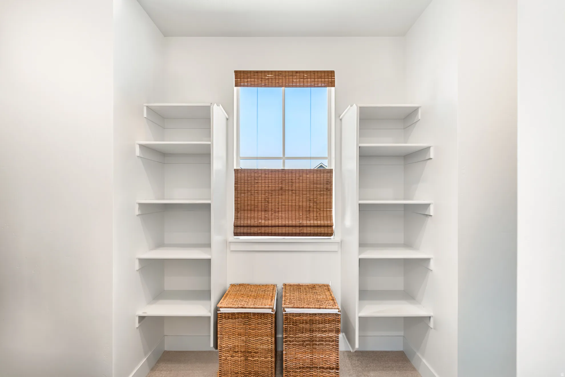 Spacious walk-in closet featuring built-in shelving, a large window with natural woven shades, and light-toned carpeting
