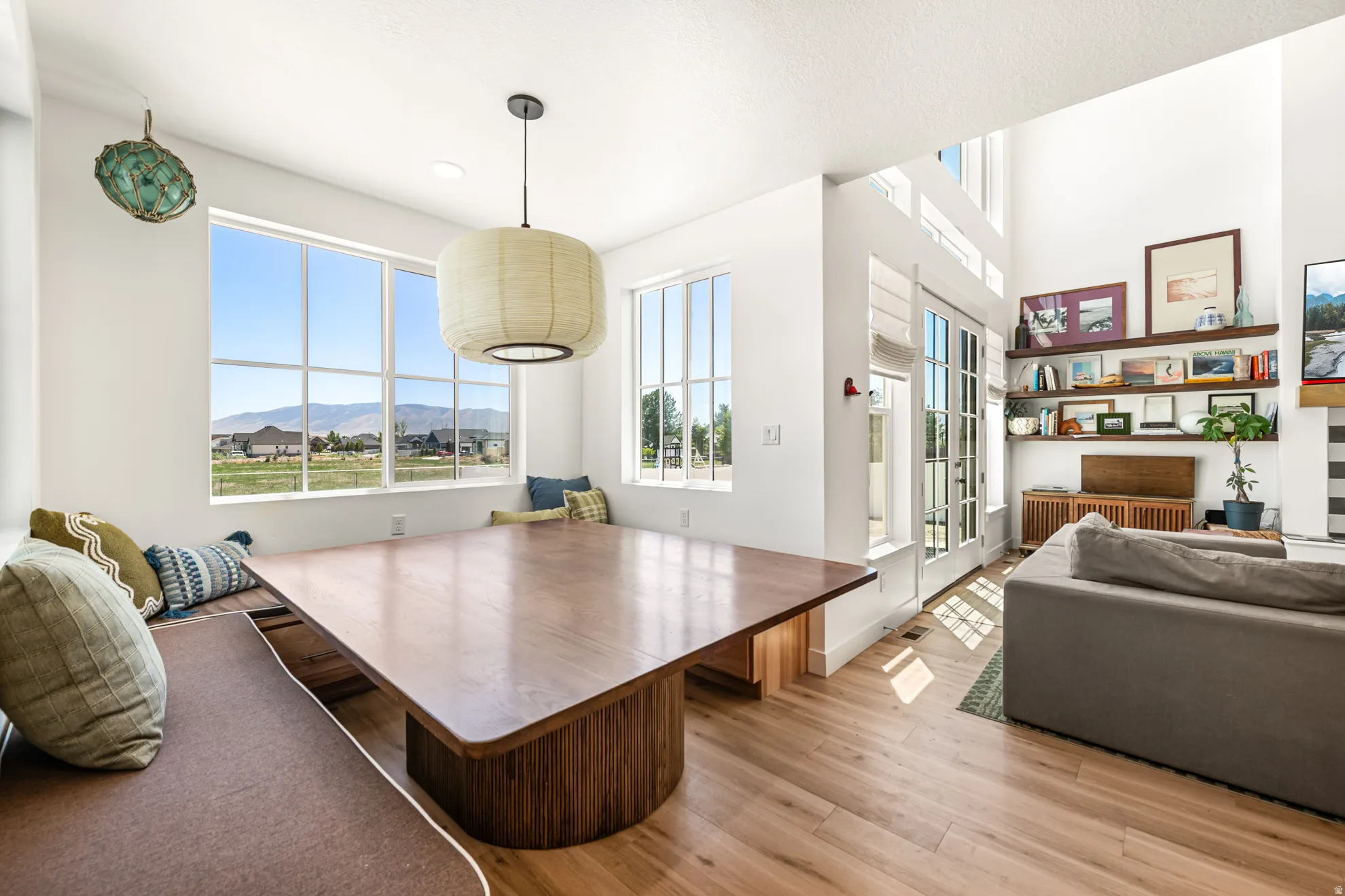 Open-concept living space with expansive windows, wood-finish flooring, a built-in dining bench, a central pendant light fixture, and double French doors