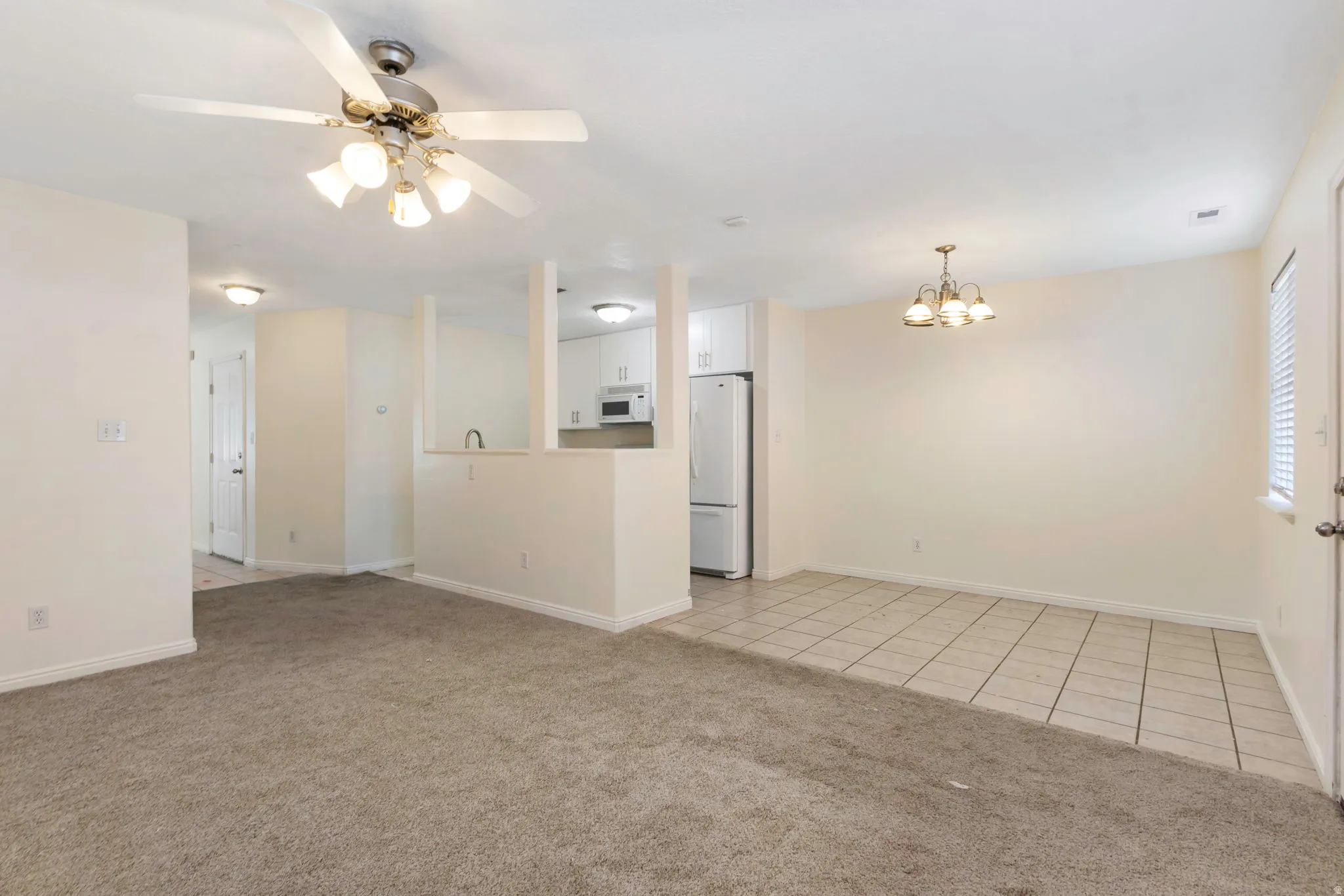 Open-concept living area featuring neutral carpeting, a ceiling fan with integrated lighting, and a dining area with tile flooring and a chandelier