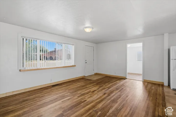 Spacious room featuring wood-finish flooring, white walls, a large window with vertical blinds, and a white ceiling-mounted light fixture