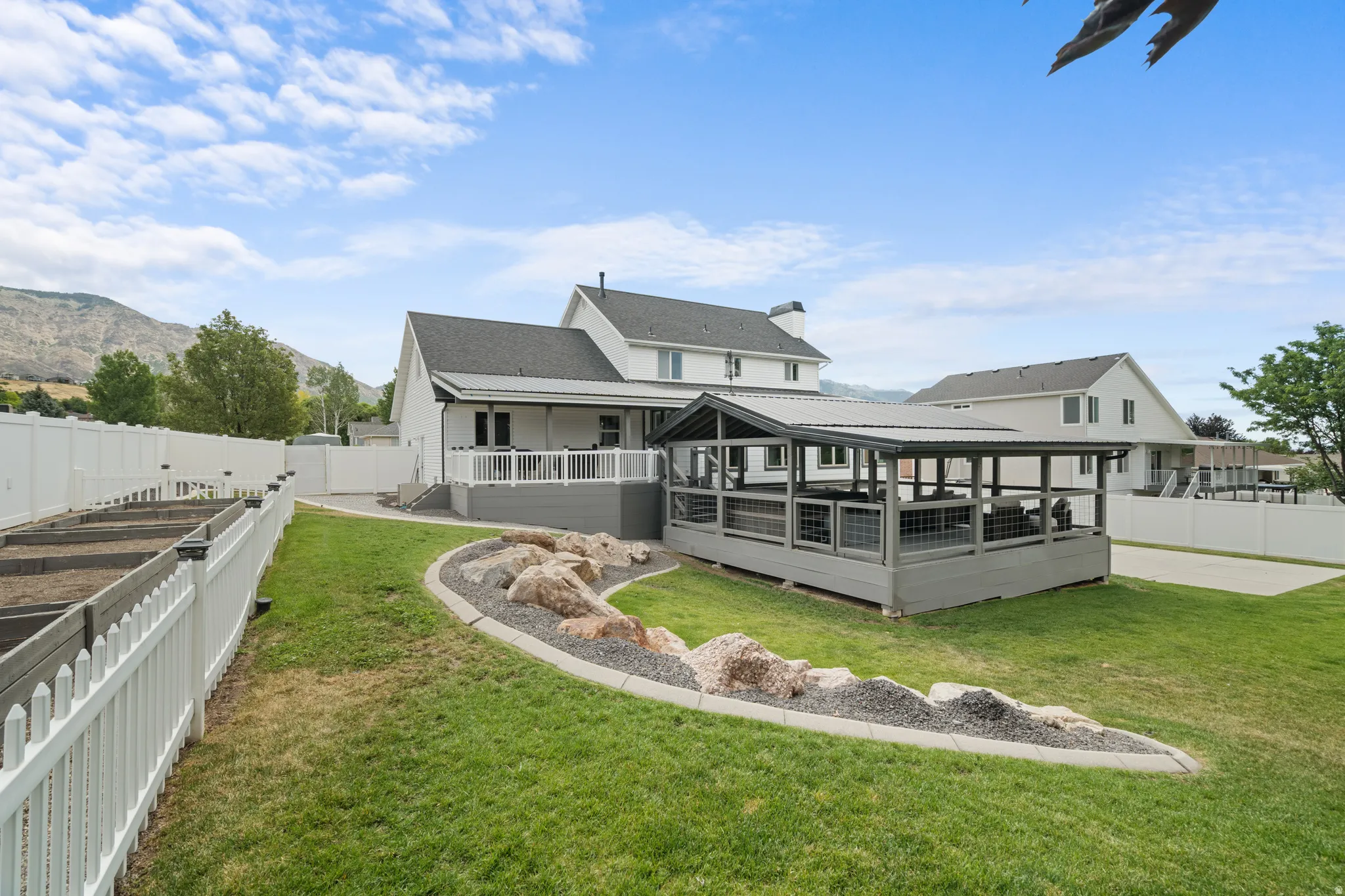 Spacious backyard featuring a covered patio, elevated deck with white railings, and a beautifully landscaped rock garden. The yard also includes eight fenced raised garden beds, providing ample space for gardening, flowers, or homegrown produce