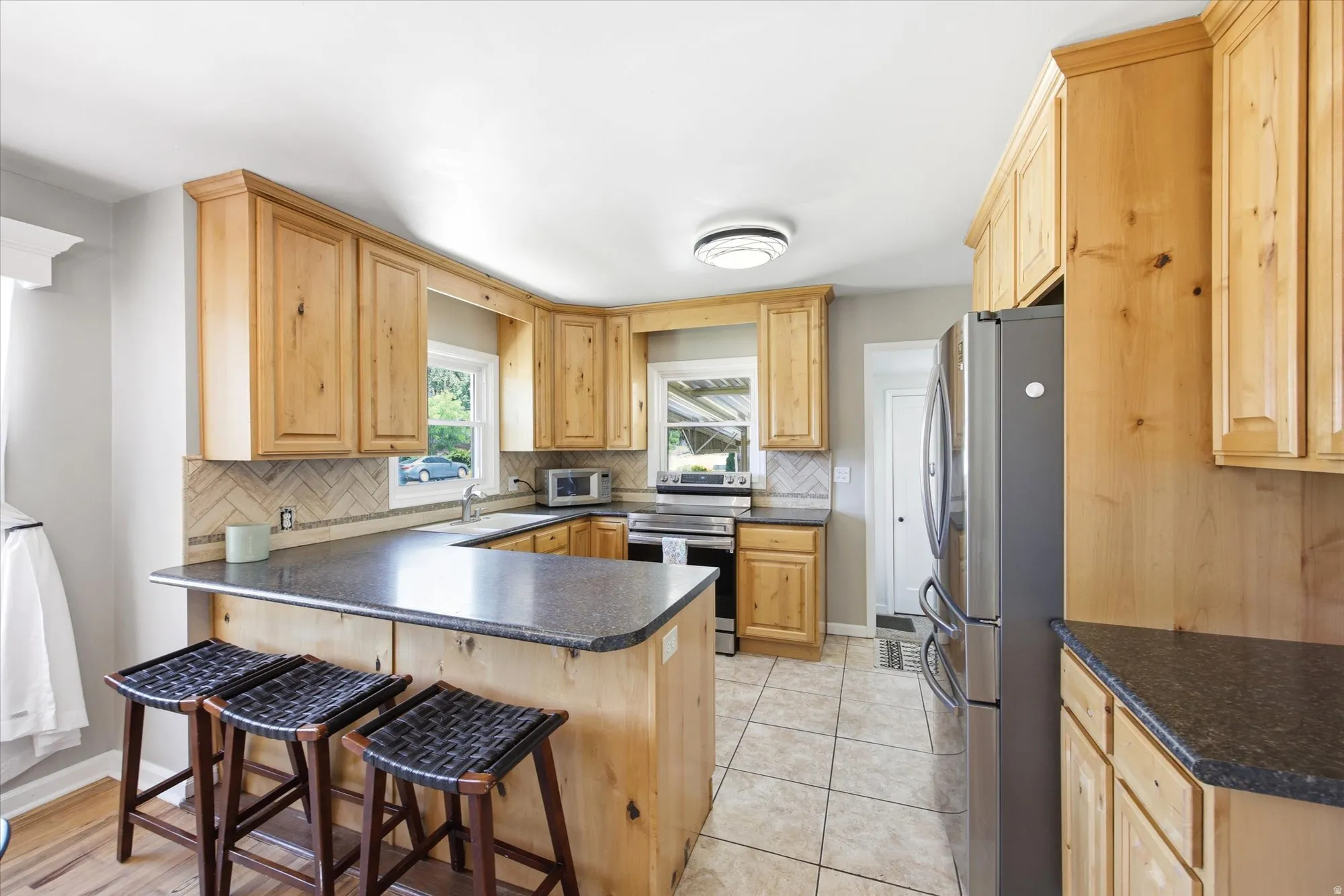 Kitchen featuring natural wood cabinetry, dark countertops, a herringbone tile backsplash, stainless steel appliances, and tile flooring