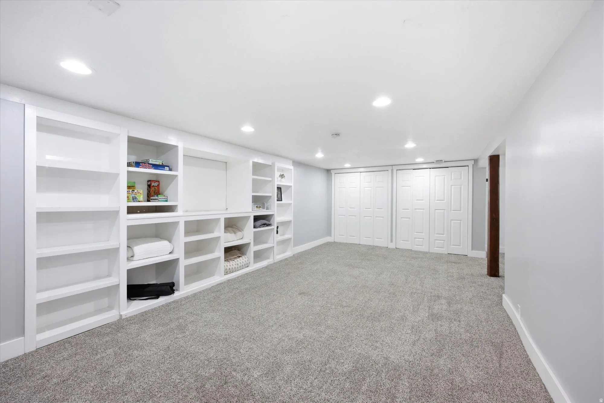 Expansive room featuring a wall of built-in white cabinetry and shelving, recessed lighting, light gray carpeting, and multiple white bi-fold closet doors