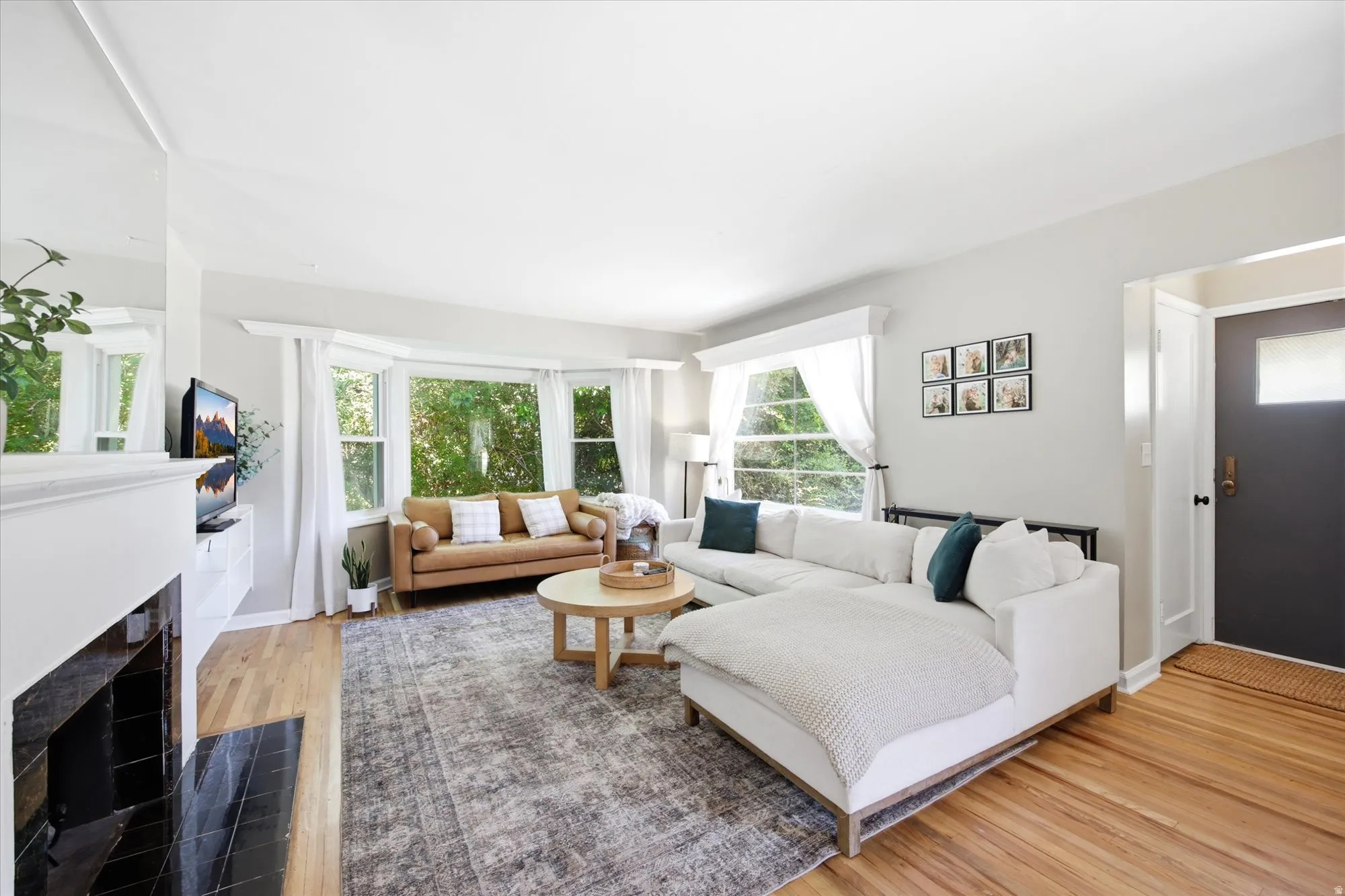 Bright living area featuring wood-finish flooring, a prominent bay window, and a white fireplace with a black tile hearth