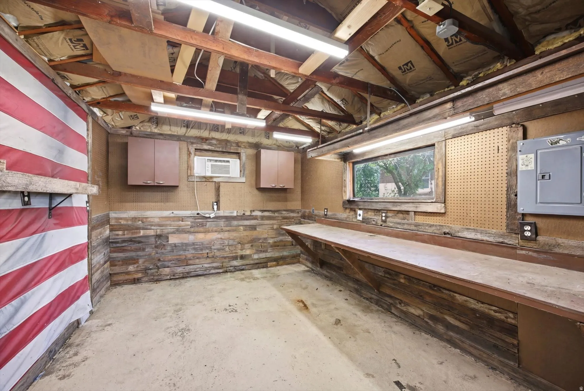 Utility room featuring a concrete floor, exposed wood beam ceiling, linear fluorescent lighting, and a built-in workbench with storage