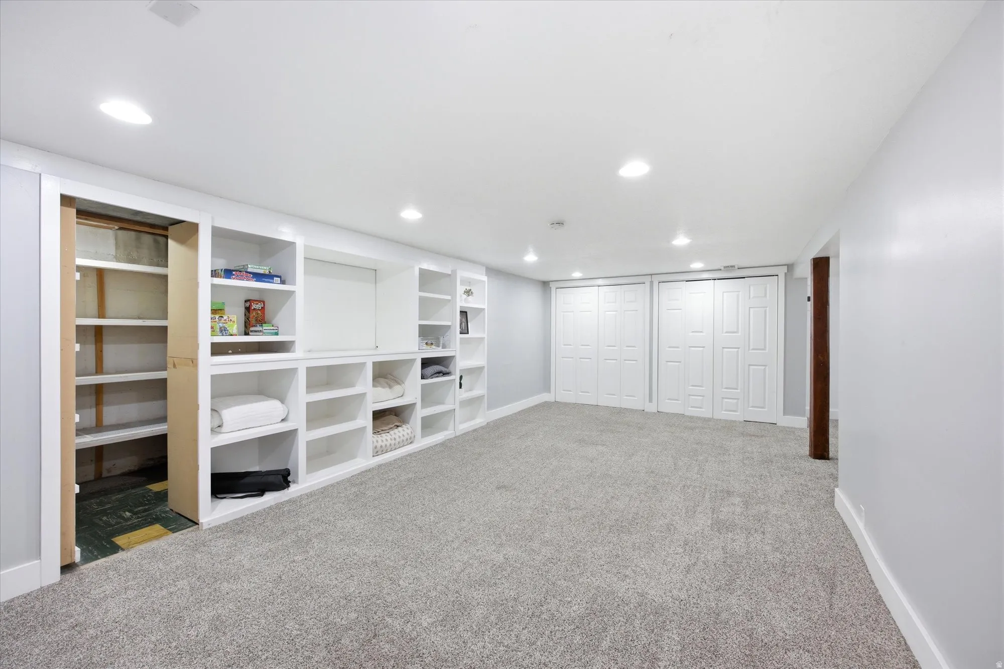 Spacious room featuring extensive built-in shelving, plush gray carpeting, recessed lighting, and white bi-fold closet doors