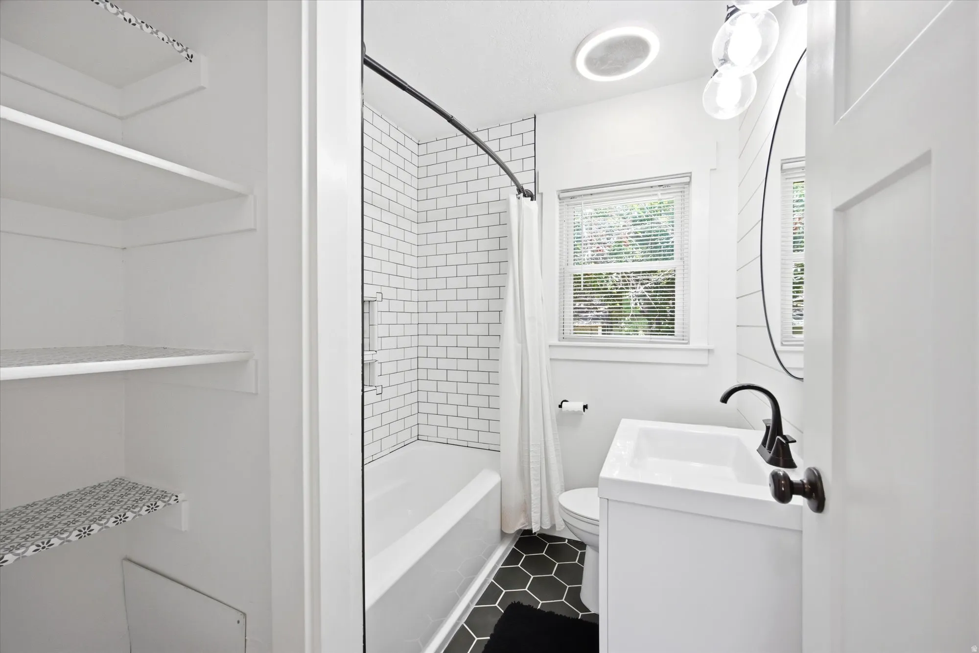 Bathroom featuring a white subway tile shower surround, dark hexagonal floor tiles, a white vanity with a rectangular sink, matte black faucet, and a round wall-mounted mirror