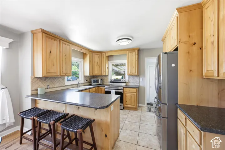 Kitchen featuring natural wood cabinetry, dark countertops, a herringbone tile backsplash, stainless steel appliances, and tile flooring