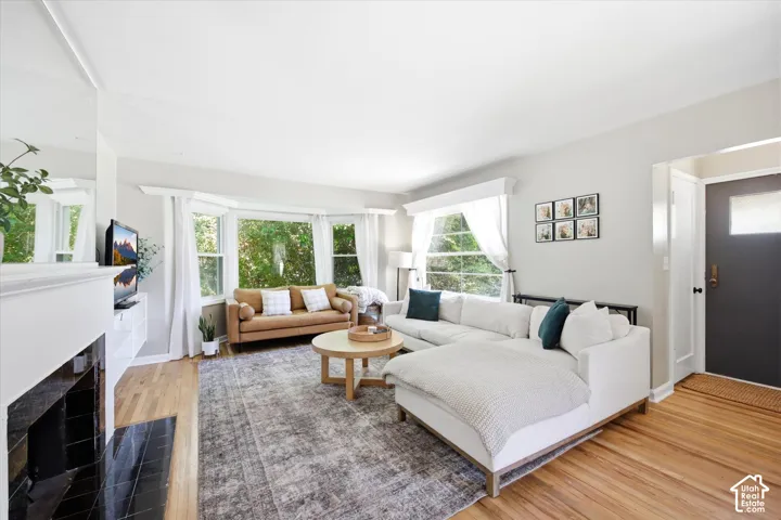 Bright living area featuring wood-finish flooring, a prominent bay window, and a white fireplace with a black tile hearth