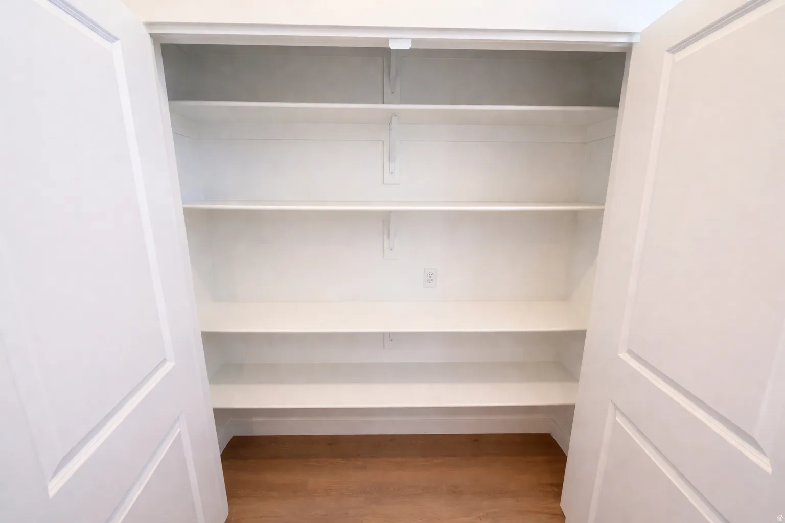 Built-in pantry with five adjustable white shelving units, a light wood-finish flooring, and white bifold doors
