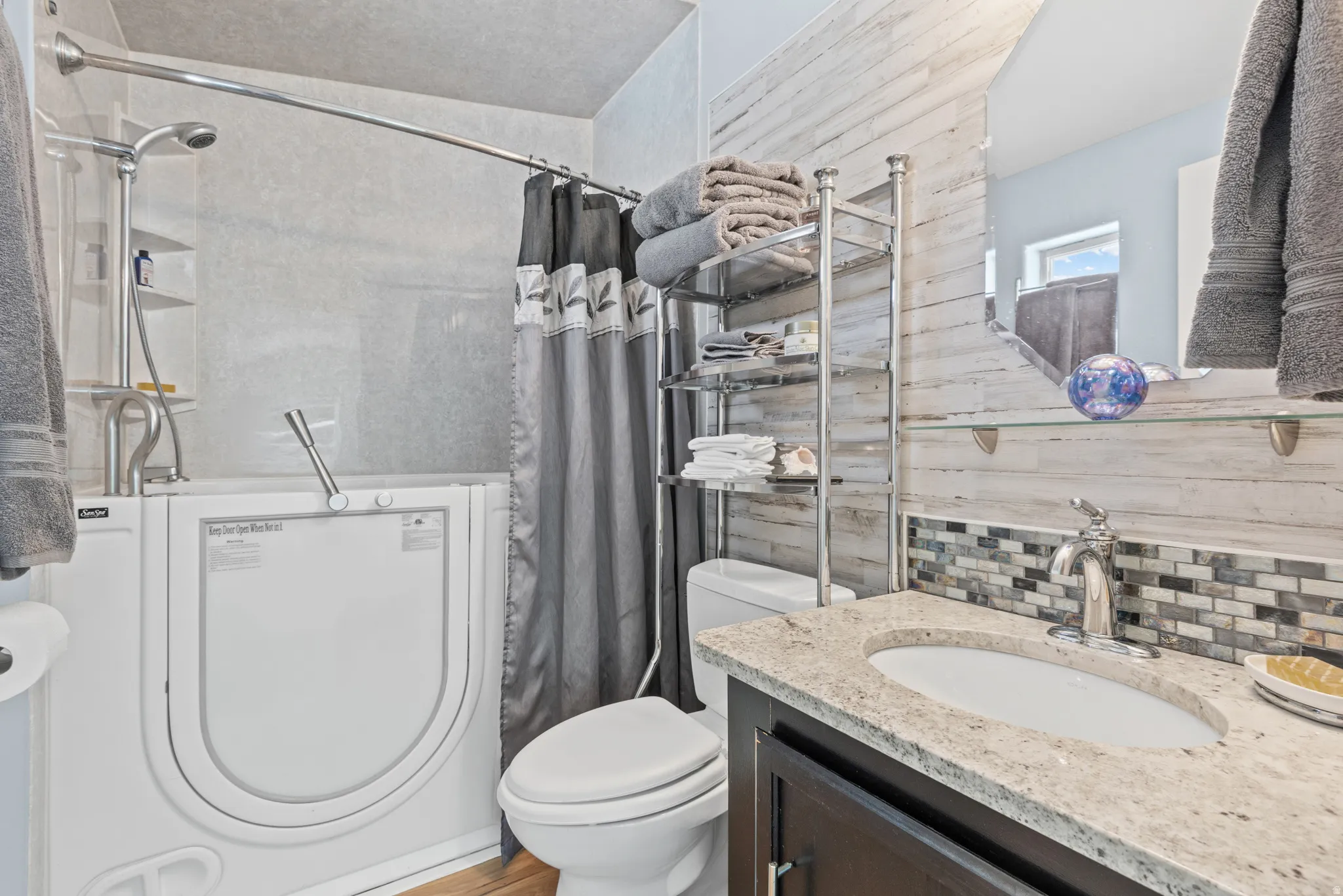 Bathroom featuring a walk-in tub with shower fixture, dark wood-finish vanity with an undermount sink and light-toned countertop, and mosaic tile backsplash