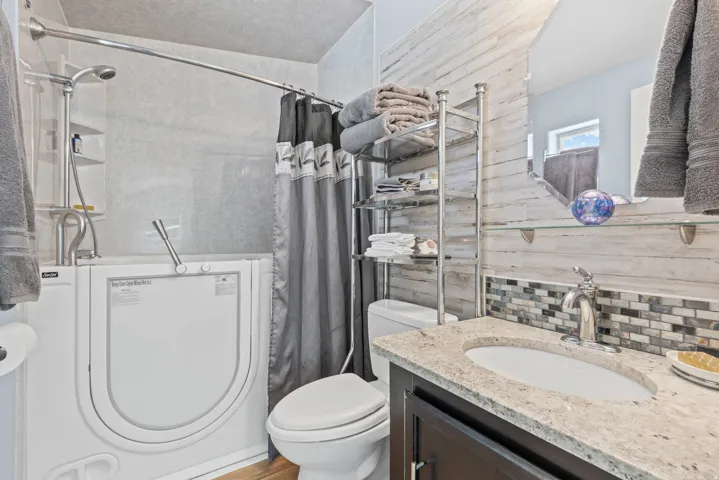 Bathroom featuring a walk-in tub with shower fixture, dark wood-finish vanity with an undermount sink and light-toned countertop, and mosaic tile backsplash