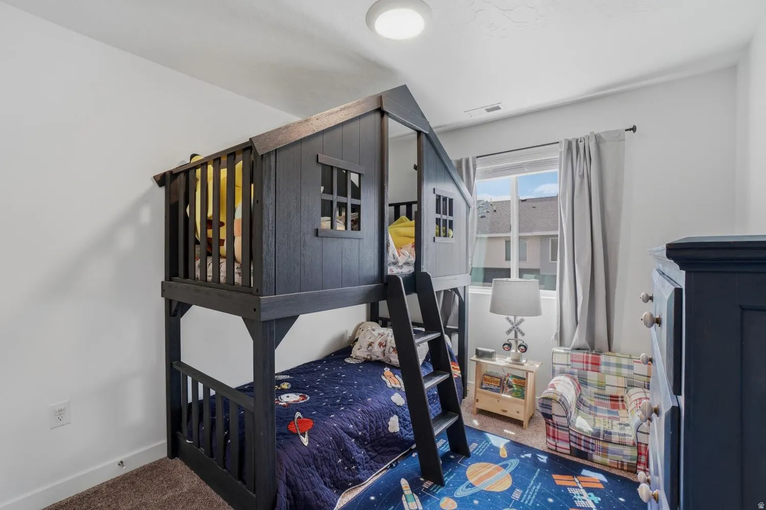 Bright room featuring a prominent built-in bunk bed with an upper-level playhouse design, white walls, a window with natural light, and plush brown carpeting
