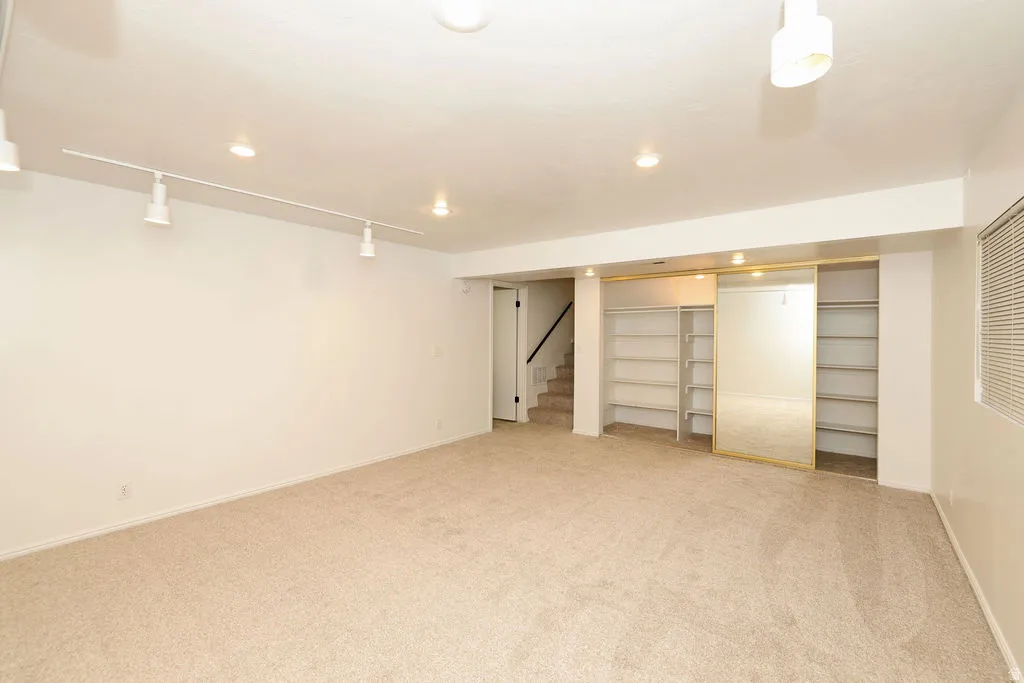 Spacious carpeted room featuring track lighting, recessed lighting, built-in shelving, mirrored sliding closet doors, and a window with horizontal blinds