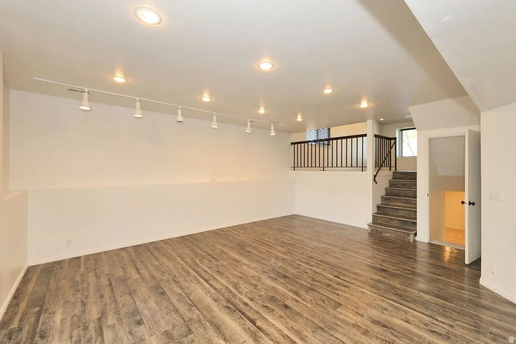 Spacious room featuring wood-finish flooring, white walls, recessed ceiling lights, track lighting, and a staircase with a black metal railing