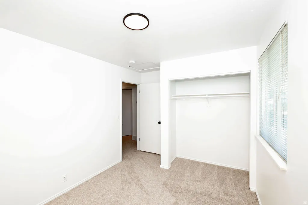 Neutral carpet flooring, white painted walls, a flush-mount ceiling light fixture, a window with blinds, and a built-in closet with shelving and a hanging rod