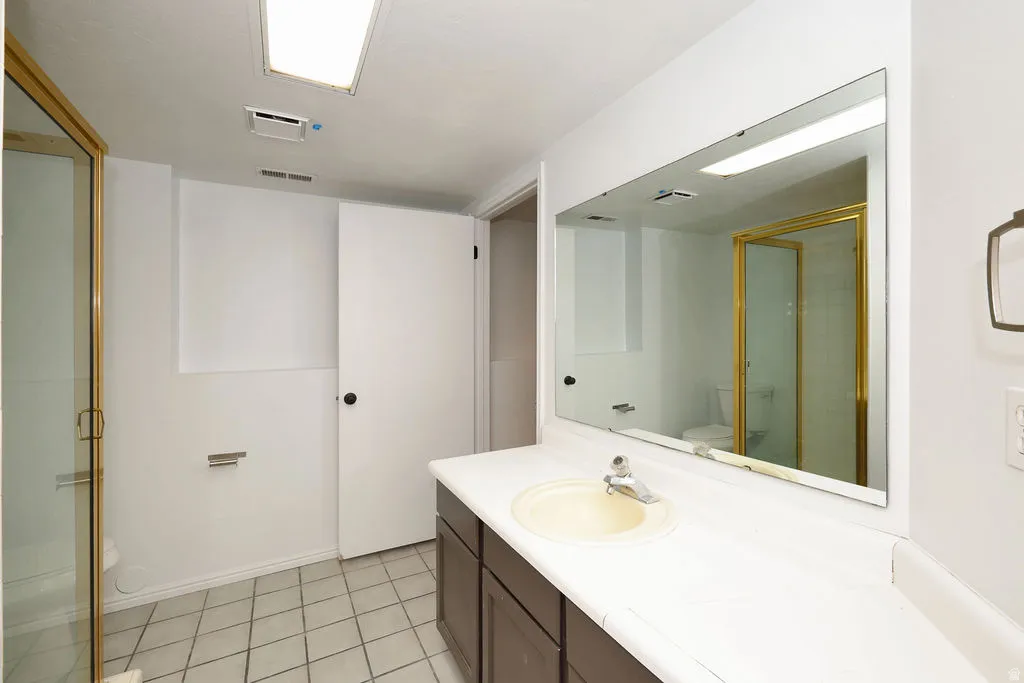 Bathroom featuring a vanity with a white countertop and integrated sink, a large wall-mounted mirror, a shower with a gold-toned frame, and light-toned tile flooring