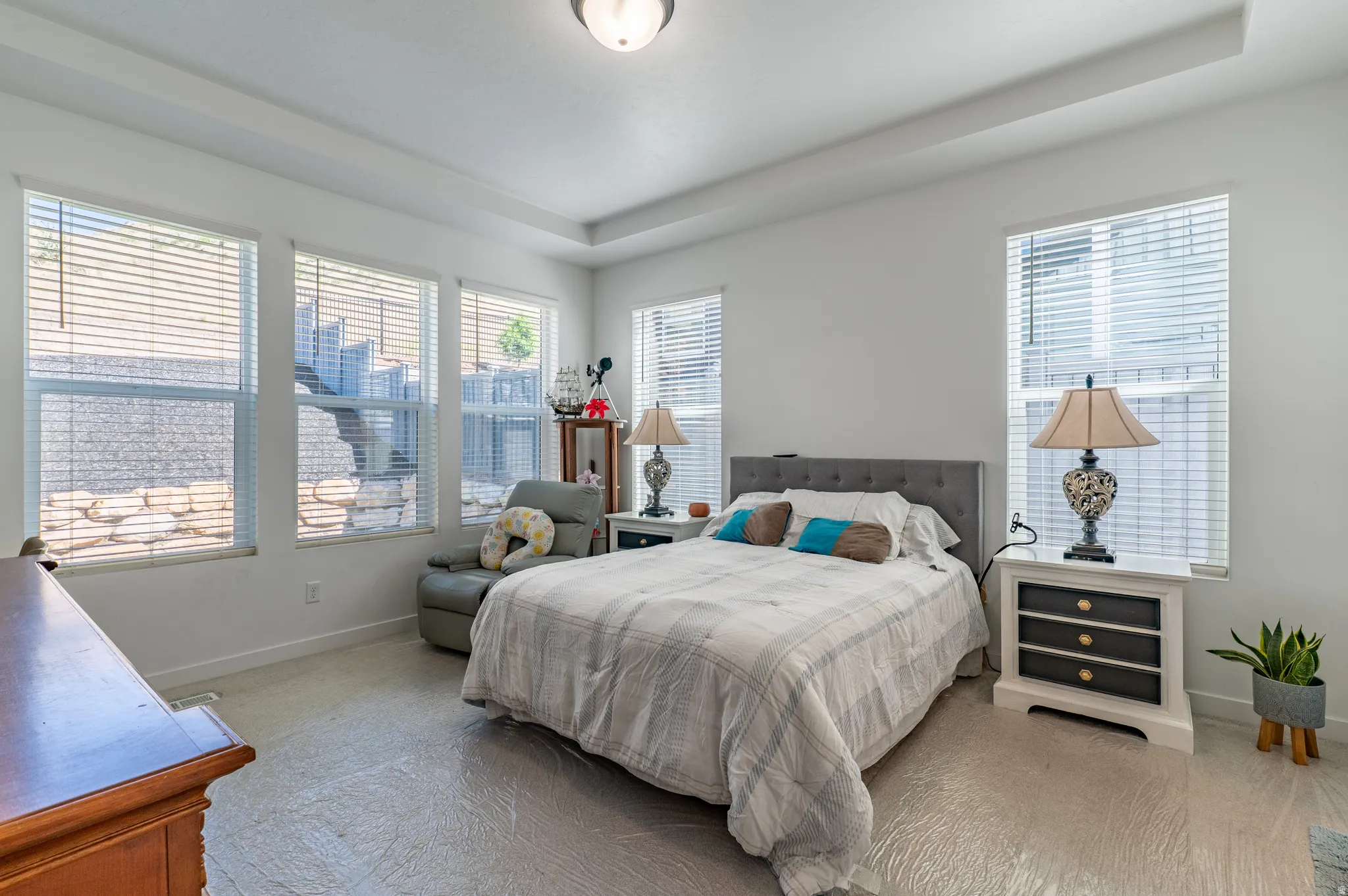 Tray ceiling bedroom featuring light-toned flooring, multiple windows with blinds, and white walls