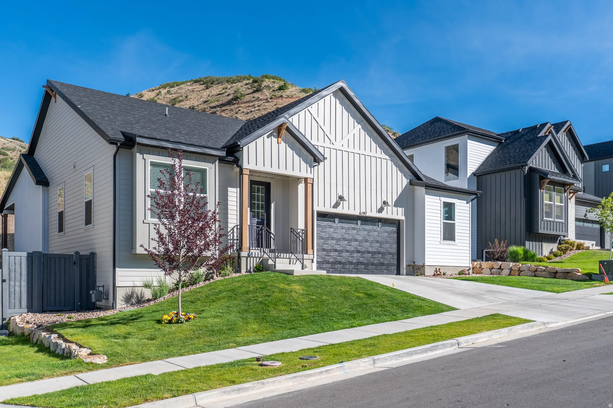 Modern farmhouse exterior featuring vertical white siding, a dark gray garage door, a covered entry with dark wood accents, and a landscaped front yard with a tree