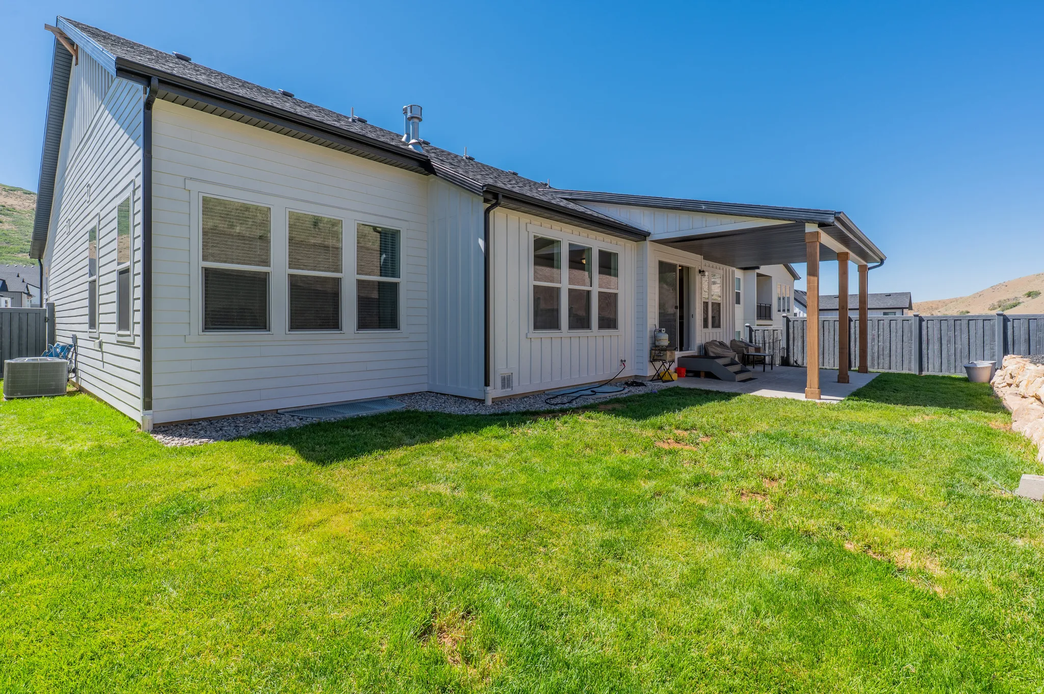 Expansive green lawn bordering a white-sided residence with black trim, featuring a covered patio with dark wood-finish posts and a concrete pad