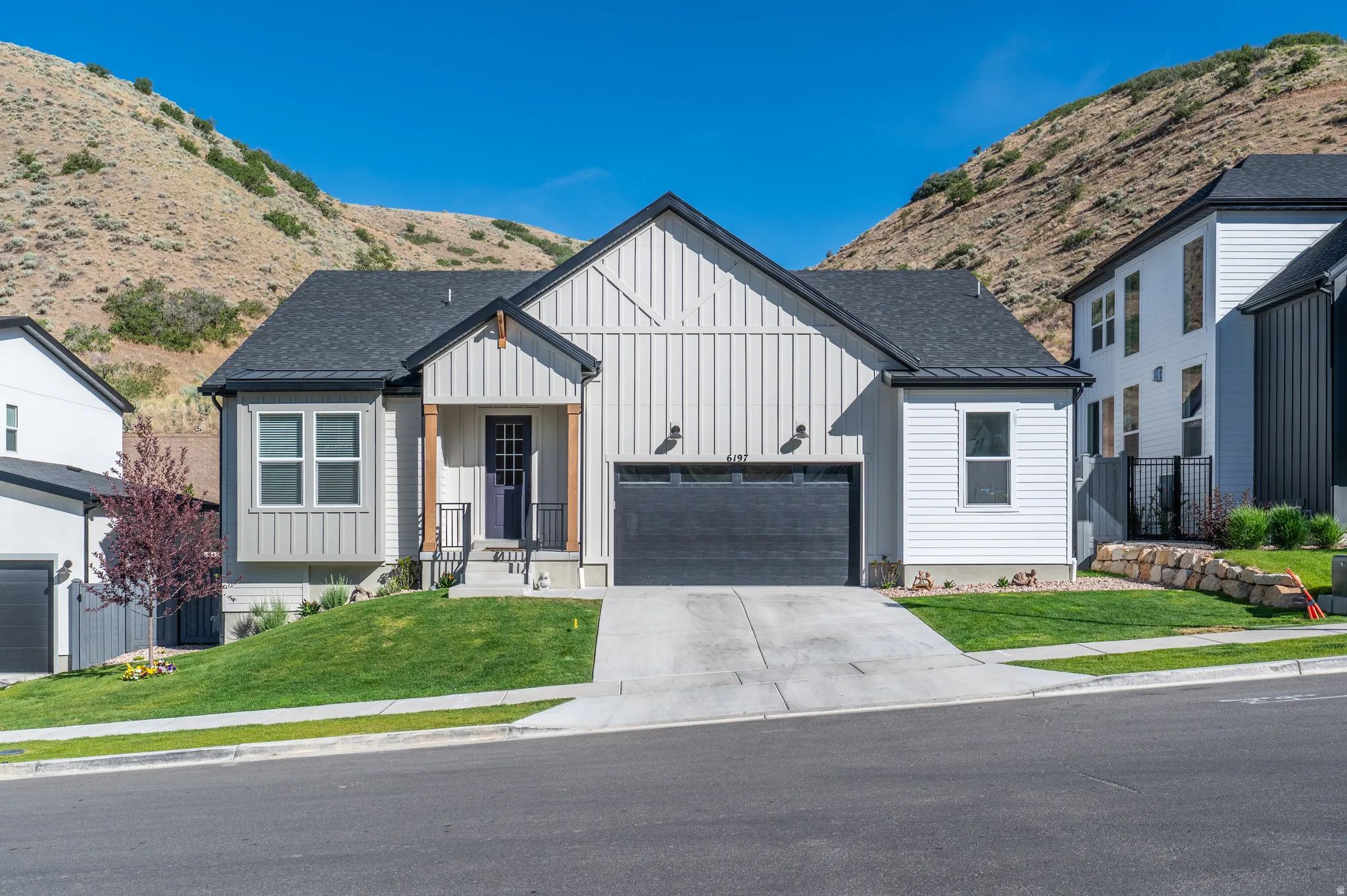 Modern farmhouse exterior featuring board and batten siding, a dark asphalt shingle roof, an integrated two-car garage, a covered front porch with wood columns, and a landscaped front yard with a retaining wall