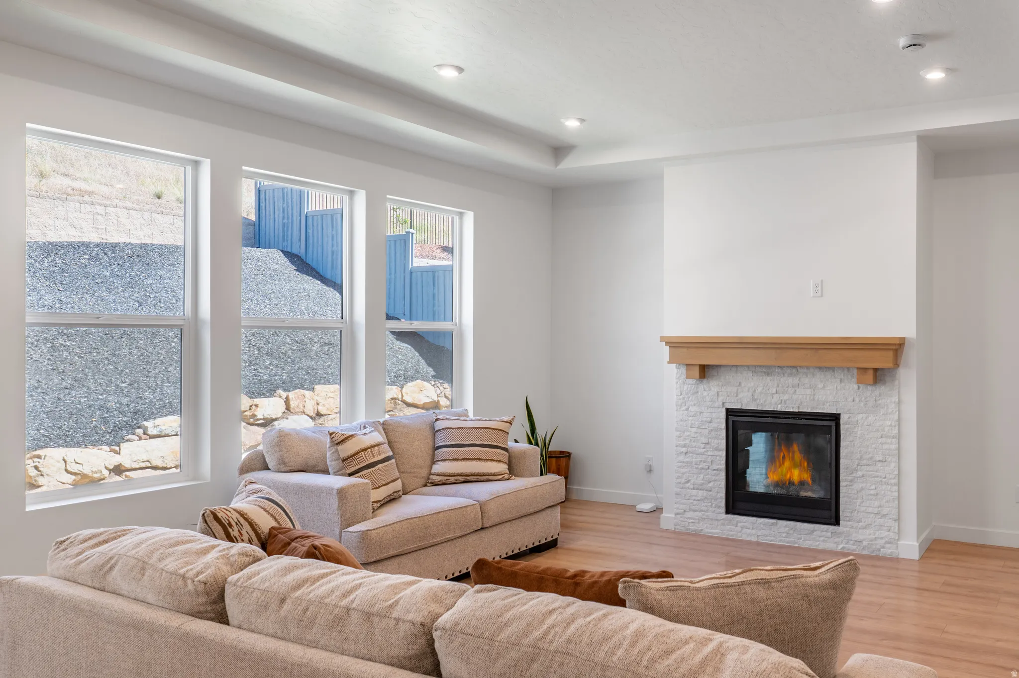 Spacious living area featuring a stacked stone fireplace with a wood mantle, recessed lighting, wood-finish flooring, and multiple windows