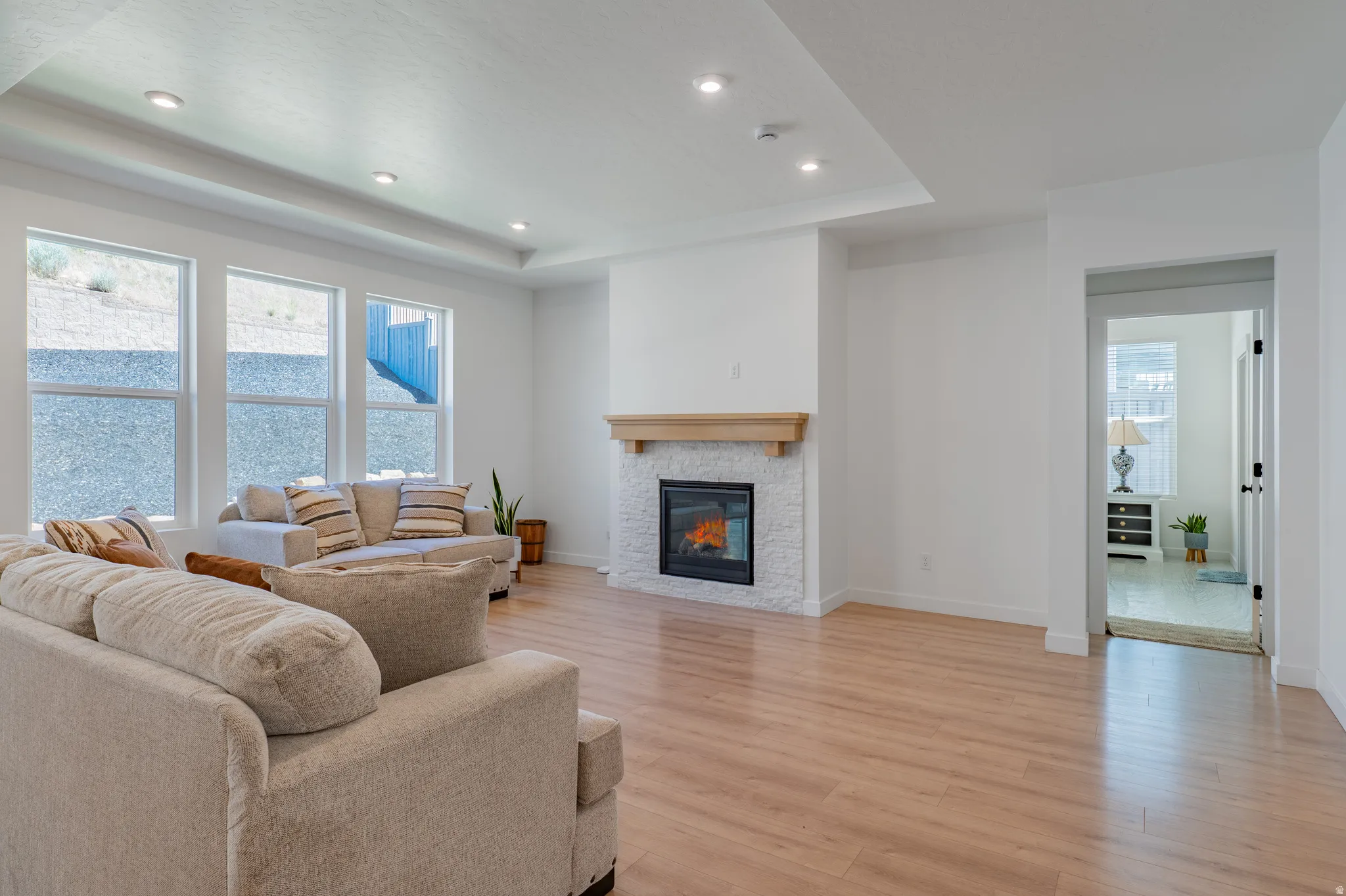 Spacious living area featuring light wood-finish flooring, a contemporary fireplace with a light-toned stone surround and wood mantel, recessed ceiling lighting, and multiple windows