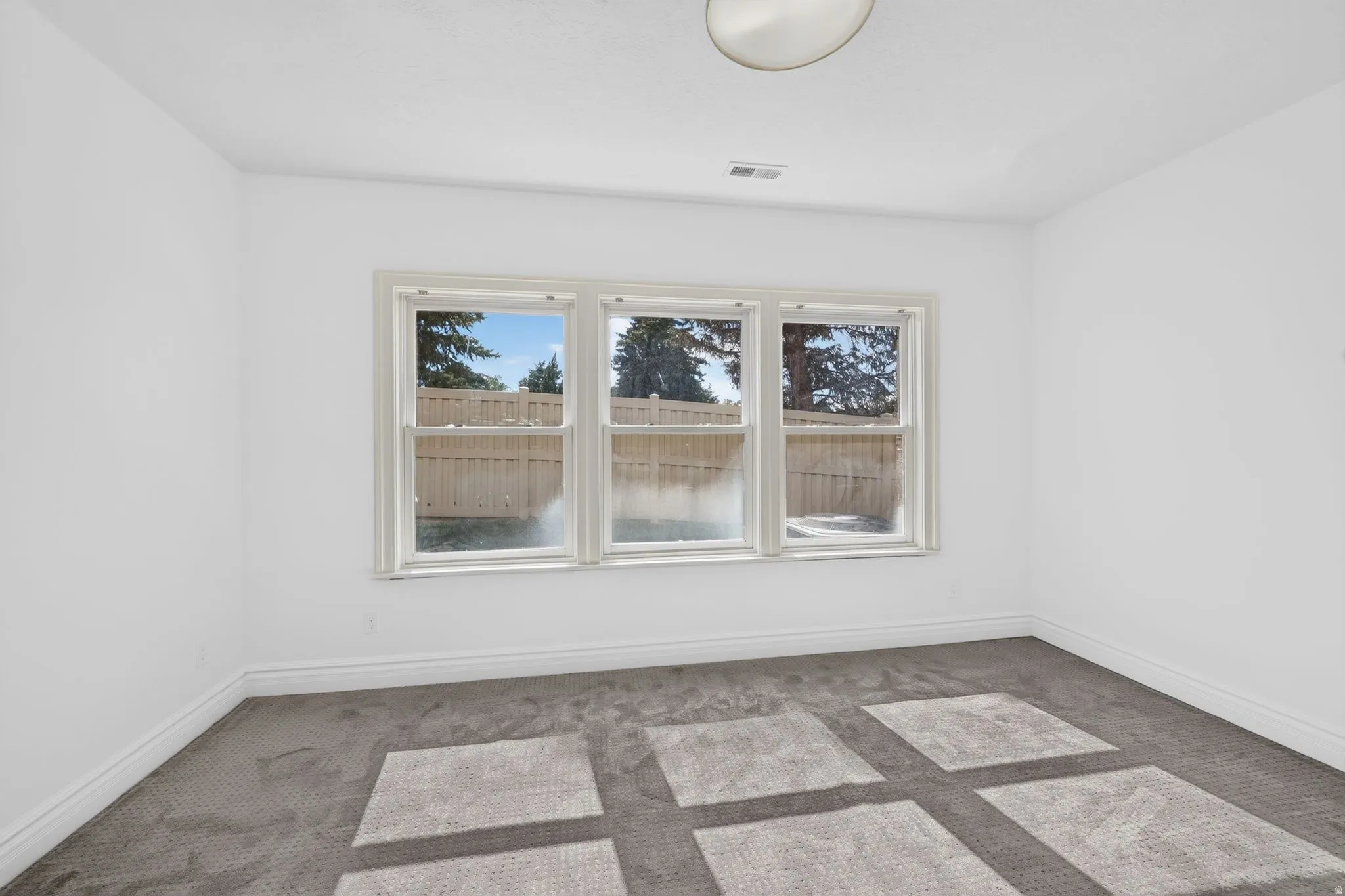 Bright room featuring three large windows, white walls, gray carpet flooring, white baseboards, and a flush-mount ceiling light