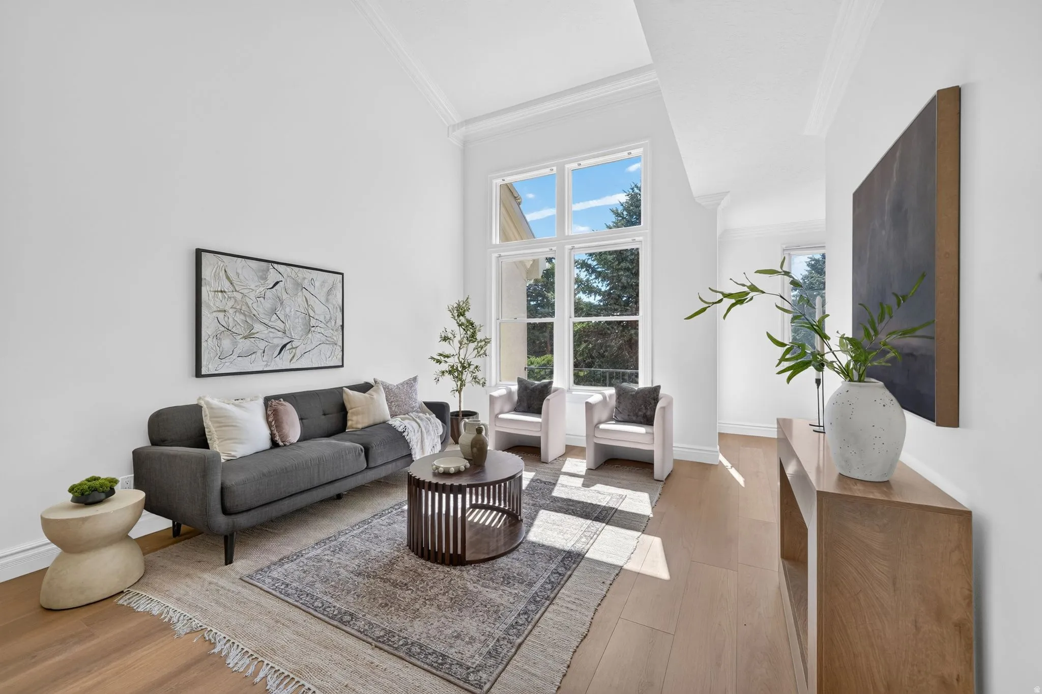 Living area featuring high vaulted ceilings, large multi-pane windows, light wood-finish flooring, white wall finishes, and crown molding