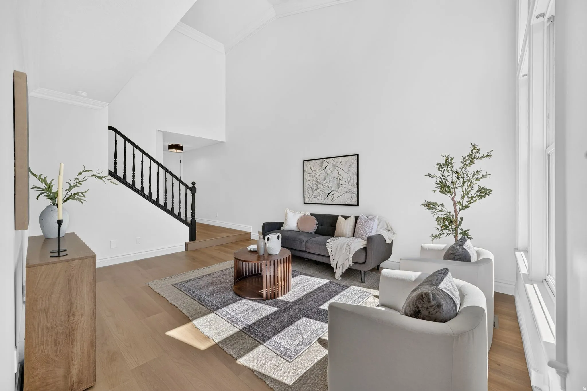 Expansive living area featuring vaulted ceilings, wood-finish flooring, a classic black spindle staircase, clean white walls, and abundant natural light from tall windows
