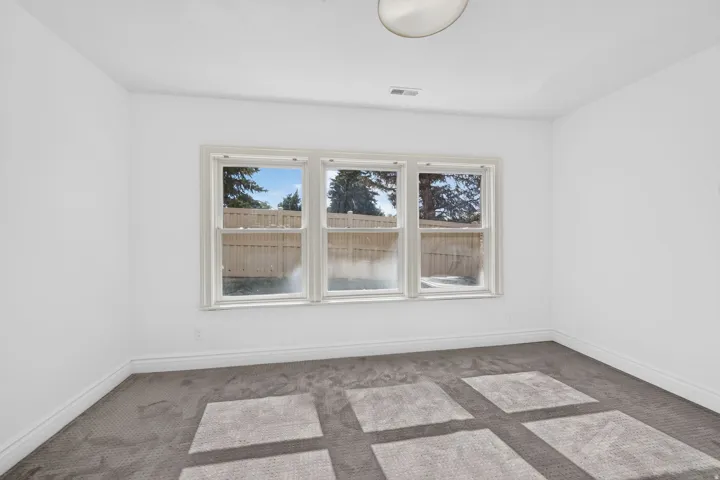 Bright room featuring three large windows, white walls, gray carpet flooring, white baseboards, and a flush-mount ceiling light
