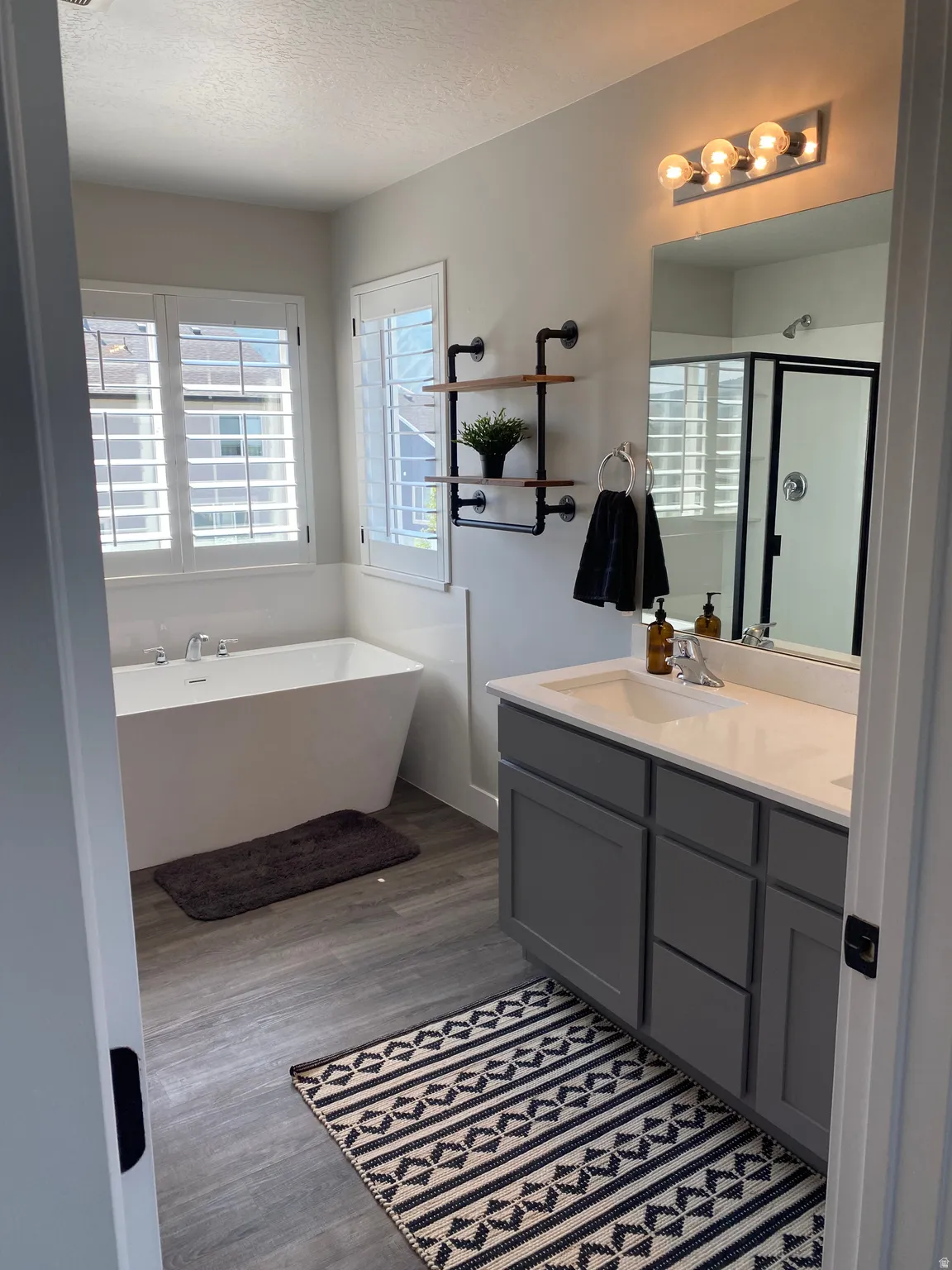 Freestanding soaking tub with white plantation shutters, dual sink vanity with gray cabinetry, and a walk-in shower with black trim