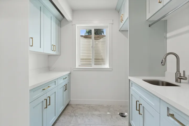 Laundry room featuring extensive light blue cabinetry with gold-tone hardware, white countertops, a stainless steel sink with a gooseneck faucet, and a window