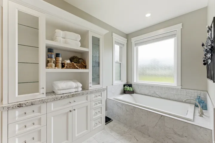 Built-in cabinetry with glass-front uppers and open shelving, integrated drop-in bathtub with marble-look surround, dual windows, recessed lighting, and a light-toned flooring