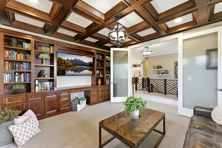 Wood-paneled coffered ceiling with recessed lighting, custom built-in cabinetry, dual glass French doors, and a decorative metal railing