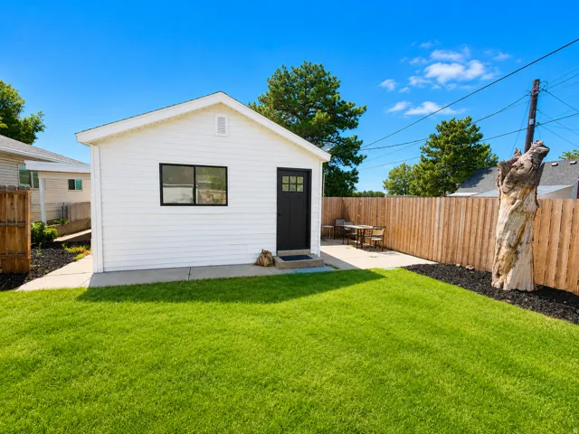 White siding structure with a black door and window, concrete patio, grass lawn, and wood privacy fence