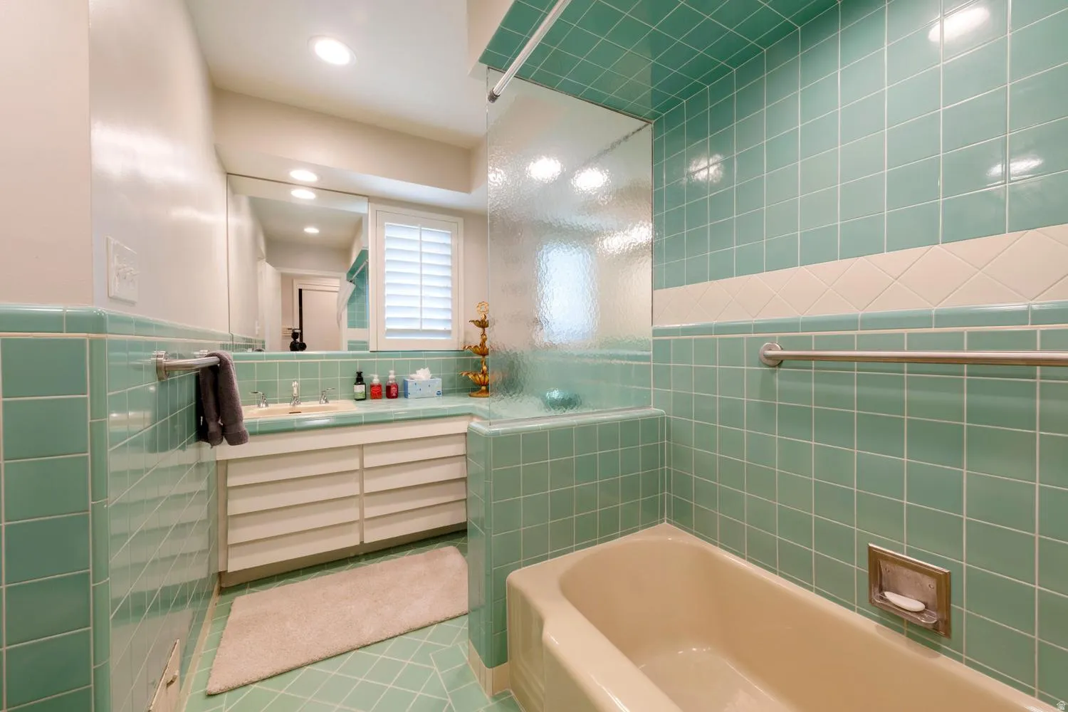 Bathroom featuring extensive teal square tilework, a built-in bathtub with a clear glass privacy screen, a vanity with louvered cabinetry, an integrated sink, and plantation shutters on the window