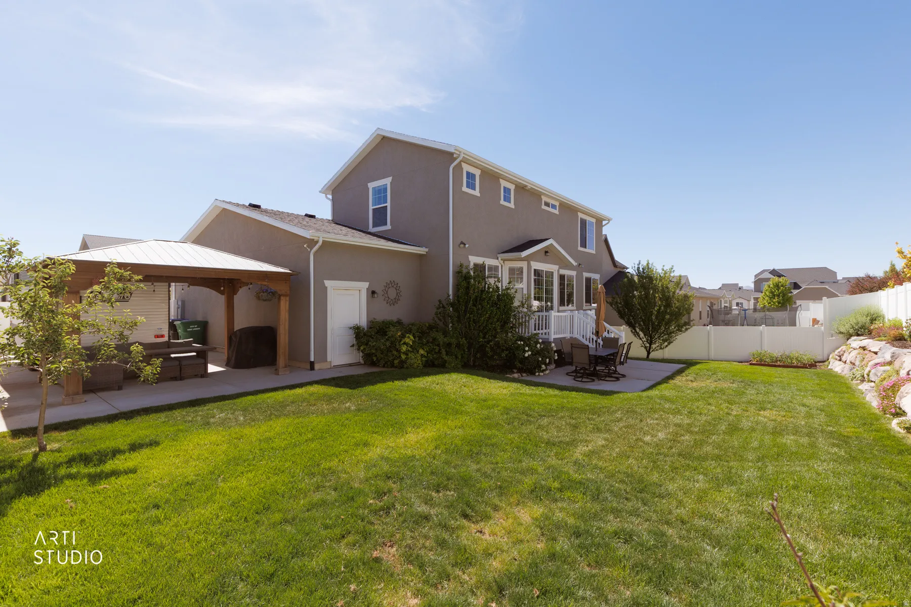 Expansive backyard featuring a manicured lawn, a covered patio structure with a metal roof, a paved patio area, an upper-level deck with white railing, and a rock retaining wall