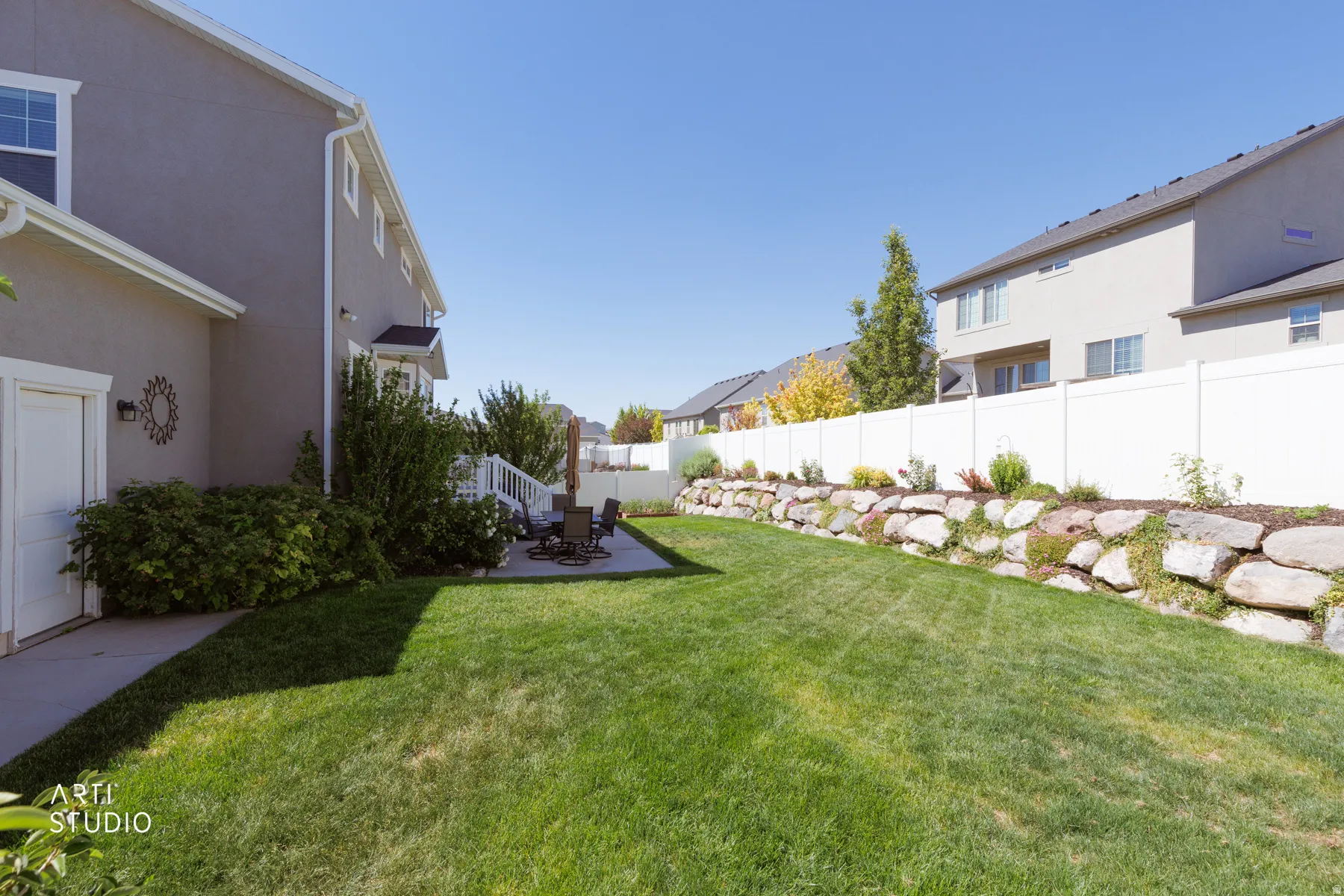 Expansive backyard featuring a stone retaining wall, mature landscaping, a white privacy fence, and a concrete patio