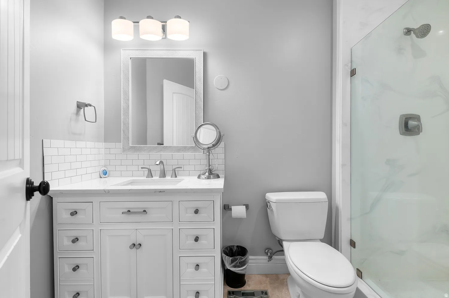 Bathroom featuring a white vanity with an integrated sink, subway tile backsplash, a wall-mounted mirror, and a glass-enclosed shower with a rainfall showerhead