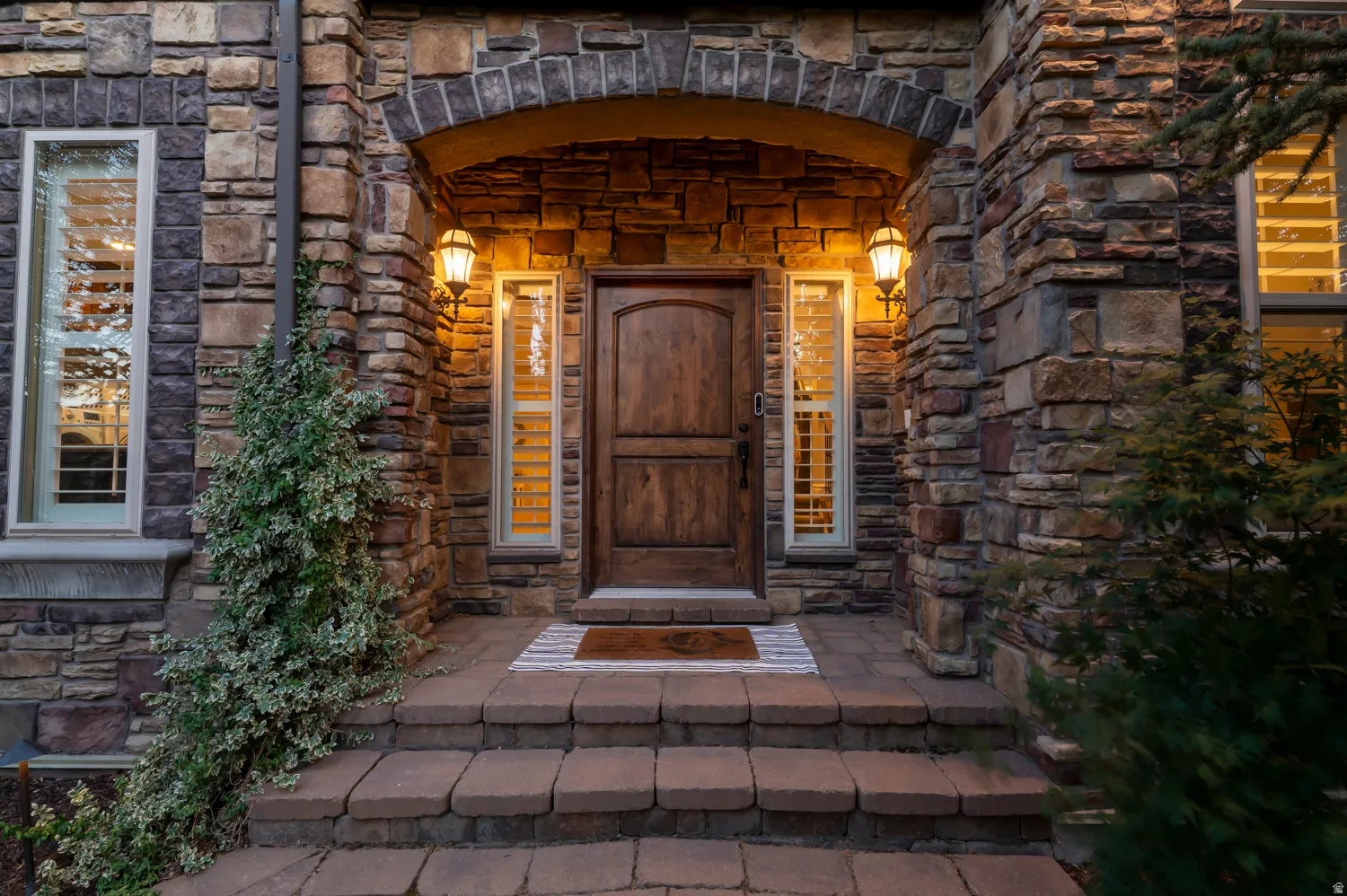 Stone facade entry featuring an arched doorway, a solid wood-finish door, and decorative sconce lighting