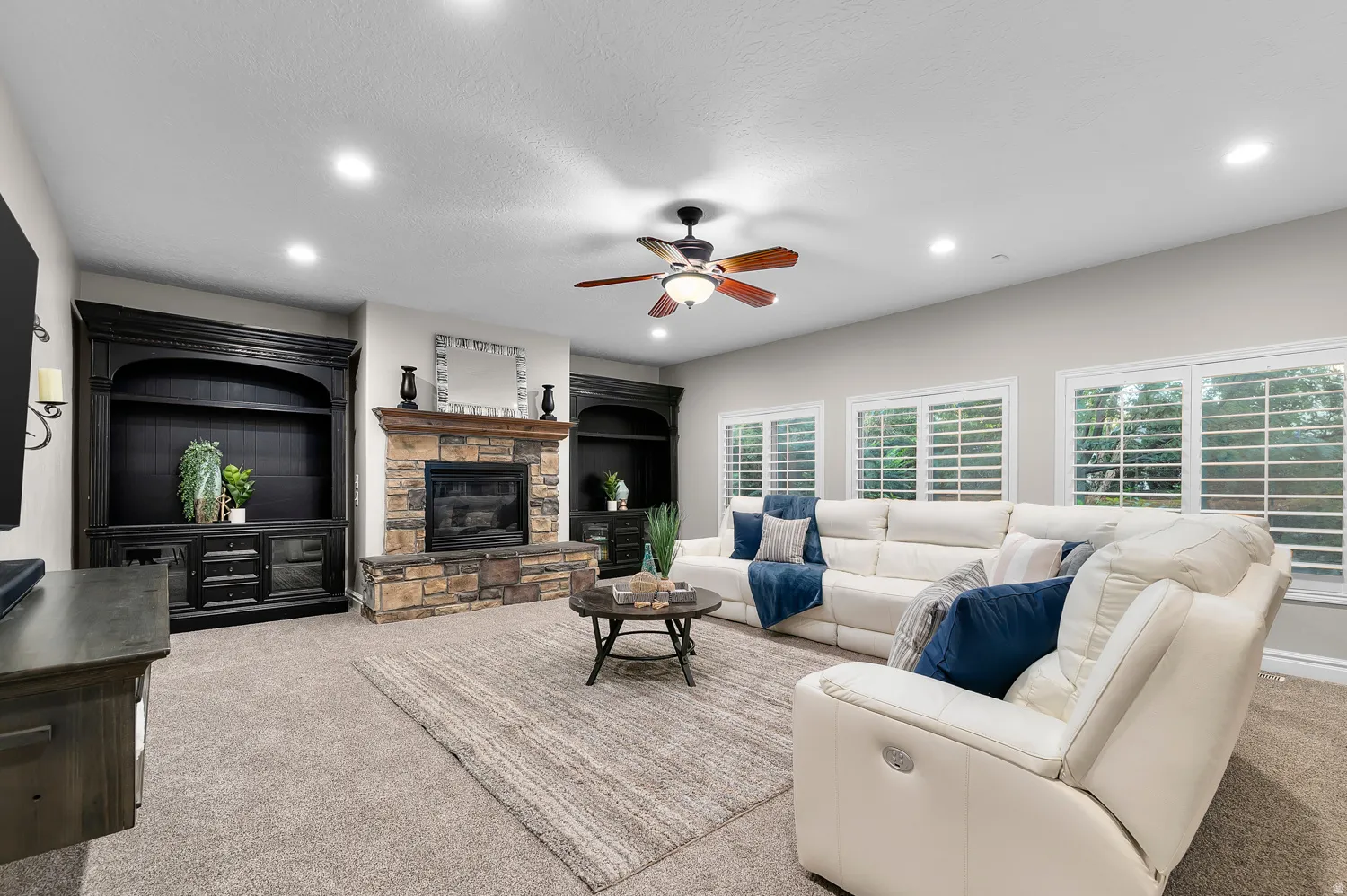 Living area featuring a stone-clad fireplace with a wood mantel, built-in cabinetry, recessed lighting, and multiple windows with plantation shutters
