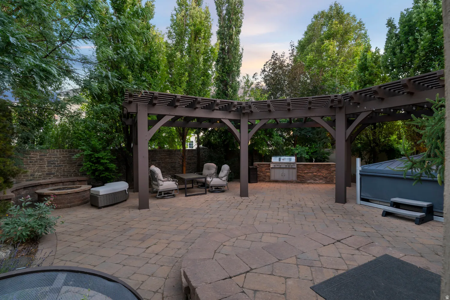 Expansive paver patio featuring a dark wood pergola, built-in outdoor kitchen with stacked stone surround, and a circular stone fire pit with integrated bench seating